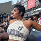 A woman seated in a stadium wearing a New York Yankees pinstripe halter crop top and matching camouflage pants, with stadium signage including Delta and Starrr in the background.