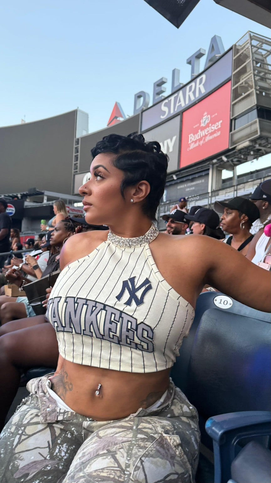 A woman seated in a stadium wearing a New York Yankees pinstripe halter crop top and matching camouflage pants, with stadium signage including Delta and Starrr in the background.