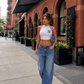 A woman walking on a city sidewalk wearing a white and navy striped halter crop top with a navy NY logo, loose blue jeans, and white boots, with a blue shoulder bag.